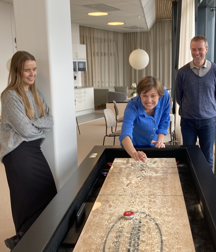 Två kvinnor och en man står vid ett shuffelboard och en av kvinnorna skickar iväg pucken.